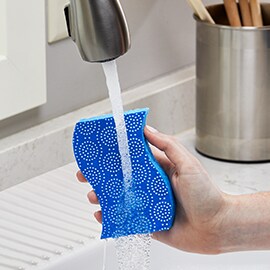 A hand holding a Scotch-Brite® Stay Fresh Non-Scratch Scrub Sponges under a running faucet.