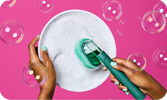 Hands washing a plate with a soapy dishwand on a pink background with bubbles.
