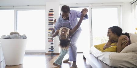 A father and daughter laughing and dancing in a modern, clean living space while the mother laughs along.