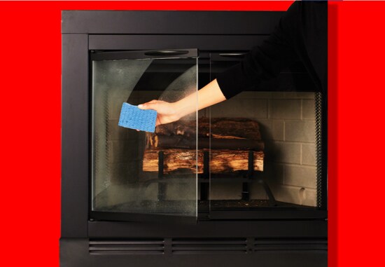 A person cleaning the door of a fireplace using a Scotch-Brite® Non-Scratch Scrub Sponge.
