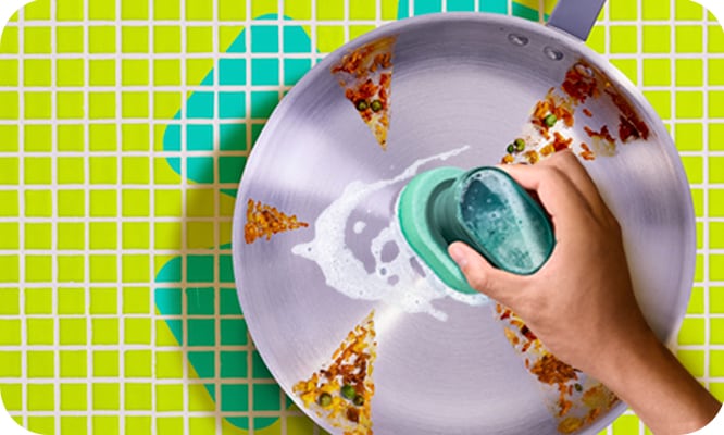 Hand scrubbing a stained frying pan, revealing its clean surface against a bright green and teal tiled background.
