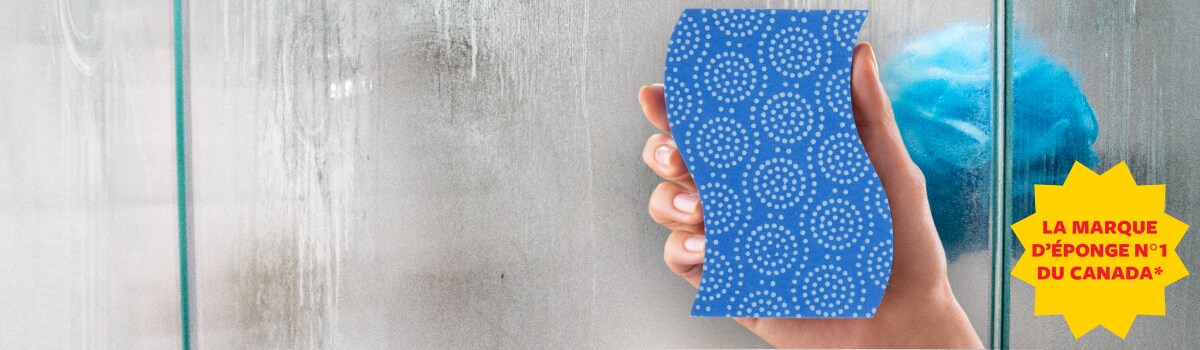 Une femme qui tient une éponge et qui s’apprête à nettoyer une douche en verre ou en acrylique.
