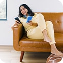 Woman in a yellow outfit sits on a brown sofa, smiling while using a smartphone, holding a sponge in her other hand.
