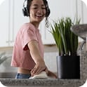 Person wearing headphones smiles while cleaning a kitchen counter near a potted plant.
