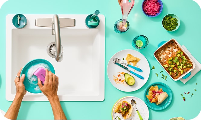 Hands washing a turquoise dish in a sink, surrounded by colorful plates, food, and drinks on a turquoise countertop.
